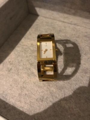 Guldfärgad klocka från Calvin Klein - Stilren guldfärgad klocka från Calvin Klein med rektangulär vit urtavla och länkarmband i metall. Klockan har minimalistisk design med diskreta visare och CK-logga på urtavlan. Perfekt accessoar för dig som gillar elegant och modern stil.