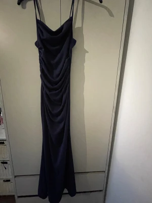 Mörkblå lång klänning med drapering - Säljer en elegant mörkblå långklänning med tunna axelband och snygg drapering framtill. Klänningen har en figurnära passform och ett mjukt, följsamt tyg som ger en lyxig känsla. Perfekt för dig som vill ha en stilren och trendig look.