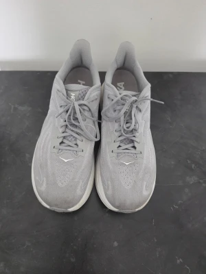 Grå Hoka sneakers med mesh och snörning - Grå sneakers från Hoka med ventilerande mesh-ovandel, klassisk snörning och en tjock, vit sula för extra komfort. Sidan har en tydlig Hoka-logga och designen är sportig och stilren. Perfekt för dig som gillar clean look och funktion.