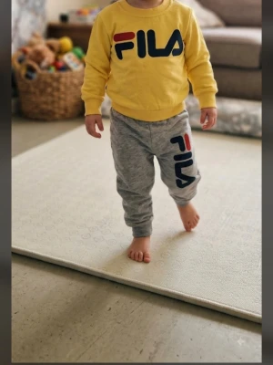  Fila set för barn 1-2år - Säljer ett sportigt tvådelat set från Fila för barn. Tröjan är gul med stor Fila-logga i svart, rött och blått framtill. Byxorna är grå med matchande logga på benet. Båda delarna är i mjukt bomullsmaterial och har bekväm passform med resår i midjan och mudd vid ärmslut och benslut.