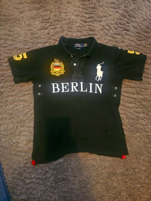 Chief keef polo - Svart pikétröja med vit BERLIN-text på bröstet, gul brodyr och stora siffror på ärmar och rygg. Klassisk krage och knappar framtill. Ikonisk logga på bröstet och detaljer i gult och rött. Perfekt för en sportig och stilren look.