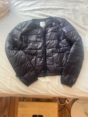 Mörkblå pufferjacka med tryckknappar - Snygg mörkblå pufferjacka med rund halsringning och tryckknappar framtill. Jackan har två sidofickor och quiltad design som ger en modern look. Perfekt för kalla dagar och enkel att matcha med olika outfits.