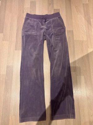 Juciy couture i Lila  - Säljer ett par lila mjukisbyxor i mjuk velour med raka ben och bred resår i midjan. Byxorna har två stora fickor fram och två fickor med knapp bak. Perfekta för chill dagar eller när du vill vara extra bekväm.