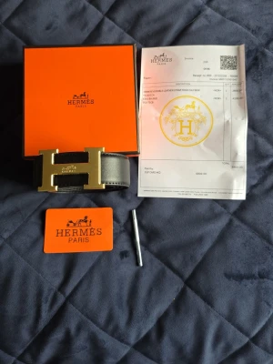 Svart Herm3s bälte med guldfärgat H-spänne - Snyggt svart bälte från Herm3s med ikoniskt H-spänne i guldfärg. Bältet är gjort i slätt skinn och kommer med originalbox och certifikat. Perfekt accessoar för att lyfta vilken outfit som helst.