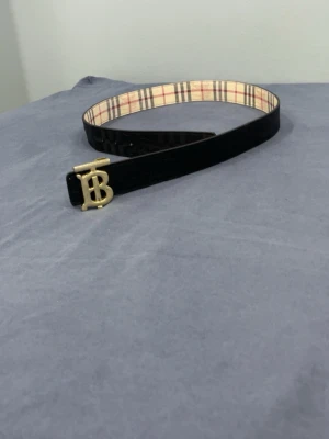 Svart bälte med guldigt spänne - Snyggt svart bälte med ett guldigt monogram-spänne och klassiskt rutigt innerfoder. Bältet har en stilren look och passar perfekt till jeans eller kostymbyxor. Insidan har ett beige, rött och svart rutmönster som ger en extra touch. Bredden är medel och materialet känns lyxigt.