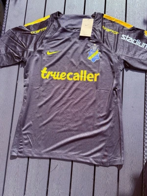 AIK matchtröja Nike svart/gul - Säljer en svart AIK matchtröja från Nike med gula detaljer och tryck. Tröjan har klubbmärke på bröstet, sponsorer som Truecaller och SVEA samt gula ränder på axlarna. Perfekt för dig som vill stötta AIK på match eller träning.
