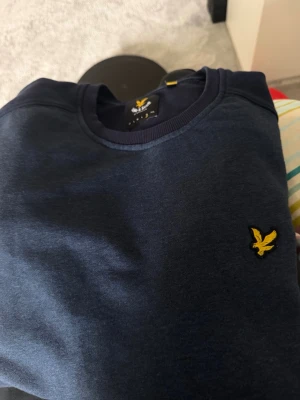 Mörkblå sweatshirt från Lyle & Scott - Snygg mörkblå sweatshirt från Lyle & Scott med klassisk rund halsringning och den ikoniska gula örnloggan broderad på bröstet. Tröjan har en enkel och stilren design, perfekt för en avslappnad look.