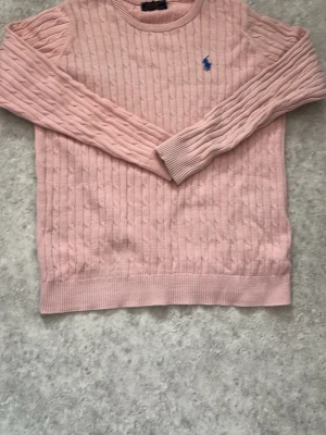 Ljusrosa kabelstickad tröja ralph lauren - Ljusrosa kabelstickad tröja från Polo Ralph Lauren med blå broderad logga på bröstet. Tröjan har rund halsringning, långa ärmar och ribbade muddar vid ärmslut och nederkant. Perfekt för en chill och stilren look.