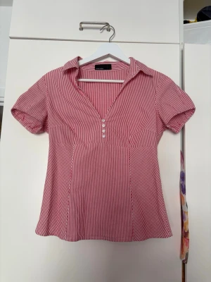 Rödvit randig blus med krage - Snygg röd och vit randig blus med korta ärmar och krage. Djup v-ringning med knappar framtill ger en trendig look. Passar perfekt till jeans eller kjol för en avslappnad men stilsäker outfit.