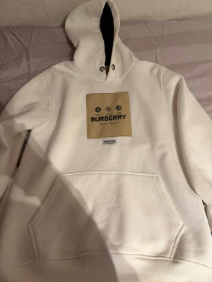 Vit hoodie från Burberry med logga - Säljer en clean vit hoodie från Burberry med stor beige patch och svart logga på bröstet. Hoodien har huva med snörning, känguruficka och ribbade muddar. Materialet känns tjockt och mjukt, perfekt för en chill och stilren look.