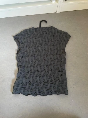 Svart mönstrad topp med struktur - Snygg svart topp med korta ärmar och ett unikt strukturerat mönster över hela plagget. Toppen har en vågig kant nertill och vid ärmslut, vilket ger en cool och modern look. Perfekt att styla med jeans eller kjol för en trendig outfit.