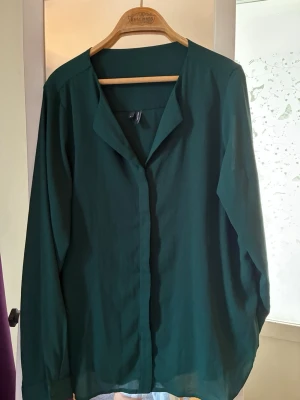 Mörkgrön blus från Vero Moda - Stilren mörkgrön blus från Vero Moda med v-ringad hals och knäppning framtill. Blusen har lång ärm och är tillverkad i ett lätt, mjukt tyg som ger ett snyggt fall. Perfekt att matcha med jeans eller kjol för en trendig look.