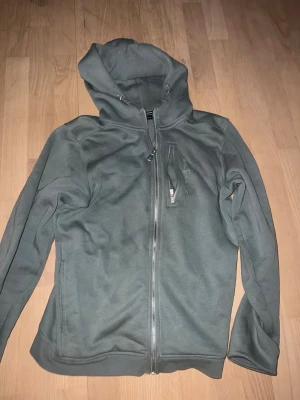 Sailracing - Säljer en stilren grå hoodie med dragkedja framtill och huva. Hoodien har en bröstficka med dragkedja och långa ärmar. Perfekt för en avslappnad look och enkel att matcha med andra plagg.