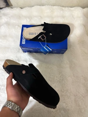 Svarta Birkenstock Boston storlek 43 - Säljer ett par svarta Birkenstock tofflor i mjuk mocka med klassisk korksula och justerbart spänne i metall. Perfekta för chill dagar hemma eller när du vill ha något bekvämt på fötterna. Levereras i originalkartong.