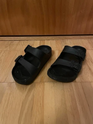 Svarta sandaler med dubbla spännen - Säljer ett par svarta sandaler med två breda remmar och justerbara spännen. Sandalerna har en klassisk slip-in design och är tillverkade i ett syntetmaterial som är lätt att hålla rent. Perfekta för en avslappnad och enkel stil.