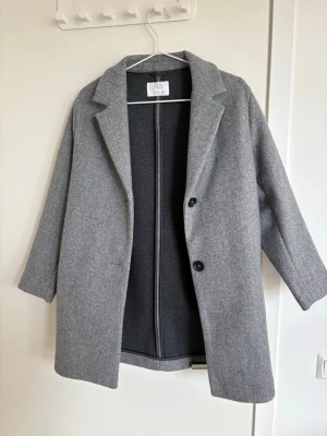 Grå kappa från Zara - Stilren grå kappa från Zara i storlek 152. Klassisk rak modell med två knappar framtill och bred krage. Kappan är tillverkad i ett mjukt material och har en minimalistisk look som passar perfekt till höst och vinter.