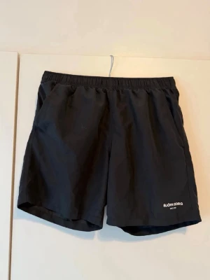 Svarta träningsshorts från Björn Borg - Snygga svarta träningsshorts från Björn Borg med elastisk midja och två sidofickor. Lätt och luftig modell med diskret logga på vänster ben. Perfekta för gymmet eller löprundan.