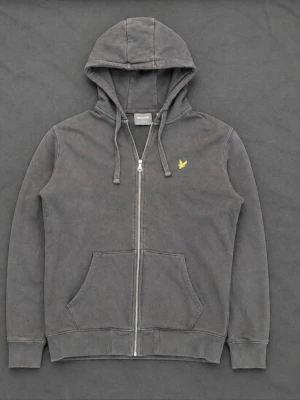 Lyle & Scott Mörkgrå Zip Hoodie - Snygg mörkgrå/tvättad svart huvtröja med hel dragkedja från Lyle & Scott. Klassisk gul örn-logga på bröstet och fickor framtill. Tillverkad i mjuk och skön bomull med ett stilrent tvättat utseende. Fint skick!  📦 Skickas snabbt! ✅ Svarar snabbt!