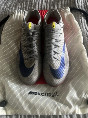 Nike Mercurial grå/blå fotbollsskor - Säljer ett par Nike Mercurial fotbollsskor i vitt med svart swoosh och gula detaljer på plösen. Skorna är i ett hyfsat skick och kan verkligen fortsättas användas. Skriv gärna om du har några funderingar och priset är ej hugget i sten och kan rabatteras vid köp av flera av mina skor!