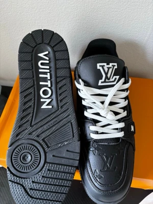 Louis Vuitton svarta sneakers - Säljer ett par svarta Louis Vuitton sneakers med vita detaljer och vita skosnören. Skorna har tydliga LV-loggor på tungan och sidan samt präglade monogramdetaljer. Tillverkade i läder med robust sula och cool streetwear-vibe.