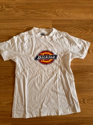 Vit Dickies t-shirt med logga - Vit t-shirt från Dickies med den klassiska färgglada loggan tryckt på bröstet. T-shirten har rund hals och korta ärmar, tillverkad i mjuk bomull för en skön och avslappnad stil.