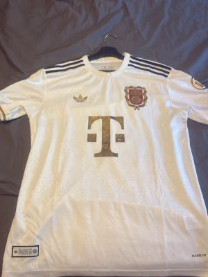 Vit Bayern München Adidas T-shirt ”Limited 125year shirt ” - Säljer en vit Bayern München T-shirt från Adidas med gulddetaljer och klubbens 125-årsjubileumsmärke på bröstet. Tröjan har svarta ränder på axlarna, rund hals och är tillverkad i ett lätt och svalt material. Perfekt för dig som gillar fotboll och snygg sportstil.