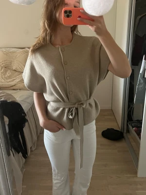 Beige kortärmad kofta med knytband - Säljer en beige kortärmad kofta med knappar framtill och ett matchande knytband i midjan. Koftan har en avslappnad passform och är tillverkad i ett mjukt stickat material. Från Zara nypris 400