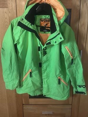 Grön vindjacka från KappAhl Woxo - Cool grön vindjacka från KappAhl Woxo med svarta detaljer och orange meshfoder. Jackan har huva, dragkedjor i kontrastfärg och flera fickor framtill. Perfekt för dig som vill sticka ut och hålla dig skyddad mot vinden.
