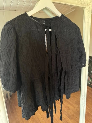 Svart puffblus från Gina Tricot - Trendig svart blus från Gina Tricot i storlek XS. Blusen har puffiga trekvartsärmar, knytband framtill och ett snyggt, strukturerat tyg som ger en cool look. Perfekt att styla med jeans eller kjol för en modern outfit.