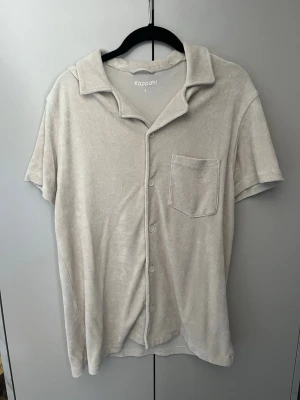 Beige frotte piké - Mjuk och skön beige frotte piké från Kappahl i frottéliknande material. Skjortan har korta ärmar, klassisk krage, bröstficka och knäppning framtill. Perfekt för avslappnade dagar hemma. Fint skick på den!