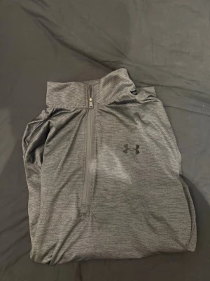 Grå långärmad träningströja Under Armour - Snygg grå långärmad träningströja från Under Armour med halv dragkedja framtill och diskret logga på bröstet. Tillverkad i ett lätt och stretchigt funktionsmaterial som andas, perfekt för träning eller löpning. Aldrig använd endast testad!
