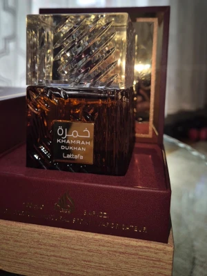 Khamrah Dukhan parfym 100ml - Khamrah Dukhan från Lattafa är en lyxig parfym i en elegant, fyrkantig glasflaska med brunt och guldigt mönster. Flaskan har en detaljerad, transparent kork och levereras i en exklusiv ask. Innehåller 100 ml Eau de Parfum.