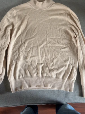 Beige polotröja från Sandro - Snygg beige stickad polotröja från Sandro. Tröjan har en hög krage och ribbade muddar vid ärmslut och nederkant. Tillverkad i mjukt material som ger en clean och stilren look. Perfekt att matcha med jeans.