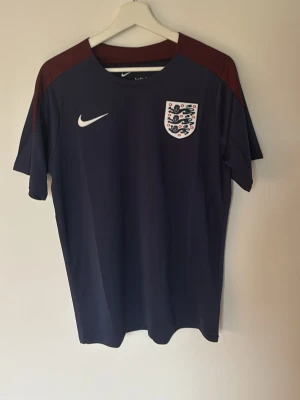 Fotbollsett england  - Snygg mörkblå fotbollströja från Nike med Englands landslagsmärke på bröstet och vita Nike-loggan. Tröjan har vinröda axelpartier och är tillverkad i ett lätt, snabbtorkande material. Perfekt för träning eller match!