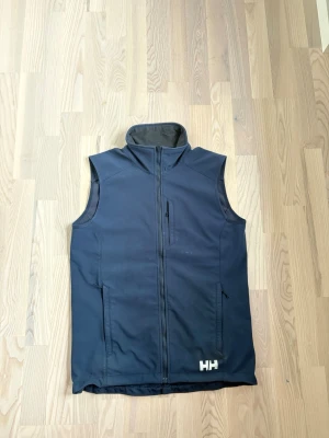Helly Hansen Väst - Navyblå väst från Helly Hansen med hög krage och dragkedja framtill. Västen har två sidofickor med dragkedja och en bröstficka. Tillverkad i ett slätt syntetmaterial, perfekt för lager-på-lager och outdoor. Storlek S men ganska stor i storleken och nypris: 1599kr.
