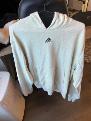 Ljusgrå hoodie från Adidas - Snygg ljusgrå hoodie från Adidas med klassisk logga broderad på bröstet. Hoodien har huva med dragsko och en stor magficka framtill. Tillverkad i mjukt bomullsmaterial som är skönt mot huden. Perfekt för en avslappnad och sportig stil.
