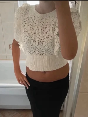 Vit stickad croppad topp med volang - Supersöt vit stickad topp med korta ärmar och volangdetaljer på axlarna. Croppad modell med ribbad nederkant och ett fint spetsliknande mönster framtill. Perfekt för dig som vill ha en trendig och luftig topp till sommare och hösten.