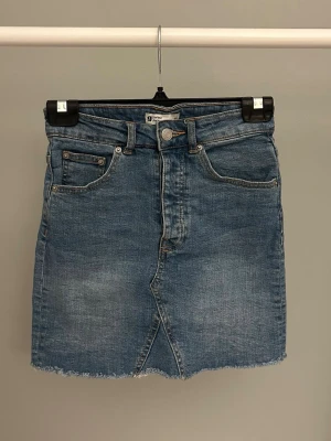 Blå jeanskjol med rå kant - Säljer en klassisk blå jeanskjol i rak modell med fem fickor och rå, fransig kant nedtill. Kjolen har knapp- och dragkedjestängning framtill samt bälteshällor. Perfekt till sneakers eller boots för en avslappnad look.
