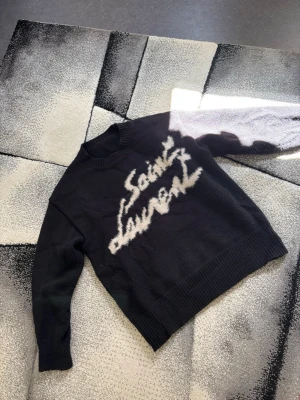 Svart stickad Saint Laurent-tröja - Säljer en svart stickad tröja från Saint Laurent med ett stort, fluffigt vitt logotyptryck diagonalt över bröstet. Fint skick. Storlek L. 