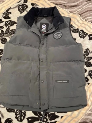 Grå dunväst från Canada Goose - Snygg grå dunväst från Canada Goose med hög krage och tryckknappar framtill. Västen har två stora fickor, diskret logga på bröstet och en patch på ena fickan. Perfekt för lager-på-lager och håller dig varm under kyliga dagar.