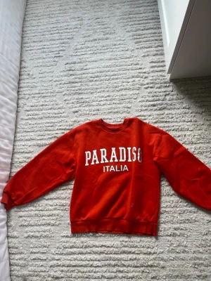 Röd sweatshirt Paradiso från Gina Tricot - Cool röd sweatshirt från Gina Tricot med texten 'PARADISO ITALIA' i vitt framtill. Tröjan har rund hals, ribbade muddar vid ärmslut och nederkant samt mjukt, borstat innerfoder. Perfekt för en avslappnad och trendig look.