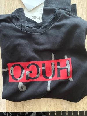 Svart hoodie med HUGO-logga - Snygg svart tröja från HUGO med stor röd och vit logga framtill. Tröjan har ribbade muddar vid ärmslut och hals, samt en klassisk avslappnad passform. Perfekt för dig som gillar streetwear och vill sticka ut med en tydlig logga.