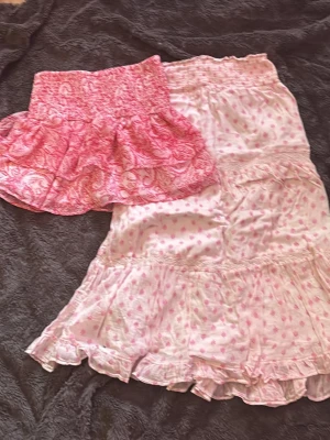 Två rosa kjolar med volang - Säljer två söta kjolar i rosa toner. Den ena är en kort volangkjol med paisleymönster och bred resår i midjan. Den andra är en lång kjol med små hjärtan och volangdetaljer. Båda är lätta och perfekta för varma dagar.