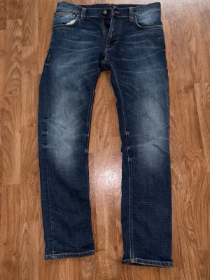 Nudie Jeans Thin Finn Indigo Dreams W32 L30 Slim Fit - Nudie Jeans “Thin Finn” i tvätten Indigo Dreams. Slim fit med smalt ben och normal midjehöjd. Mörk indigo med fina, naturligt framslitna fades på lår och bakom knäna. Storlek: W32 L30 Material: 99% ekologisk bomull, 1% elastan Dragkedjegylf, femficksmodell Skick: Mycket bra – inga hål eller fläckar. Använda, vilket syns i tvätten på ett snyggt sätt. Skickas inom 2 dagar – hör av dig vid frågor!