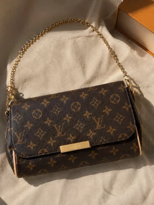 Handväska med mönster  - Snygg handväska i klassiskt brunt monogrammönster med guldiga detaljer och kedjerem. Väskan har en rektangulär form och är tillverkad i canvas med läderdetaljer. Perfekt för dig som gillar lyxiga accessoarer och tidlös design.