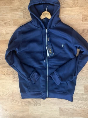 Mörkblå hoodie från Polo Ralph Lauren - Snygg mörkblå hoodie från Polo Ralph Lauren med dragkedja framtill och klassisk logga broderad på bröstet. Hoodien har huva med dragsko och är tillverkad i mjukt material som känns skönt mot huden. Perfekt för en avslappnad och stilren look.