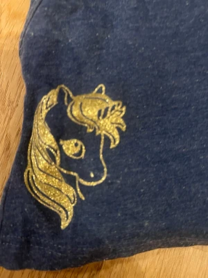 Mörkblå shorts med glittrig häst - Mörkblå shorts med resår i midjan och en glittrig guldfärgad häst på ena benet. Lite noppriga därför priset, i storlek 110-116. Skriv för mer frågor!