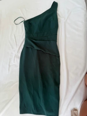 Mörkgrön oneshoulder långklänning - Stilren mörkgrön långklänning med oneshoulder-design och smal axelrem. Klänningen har en figurnära passform och är tillverkad i ett slätt tyg som ger en elegant look. Perfekt för dig som vill sticka ut med en unik och trendig siluett.