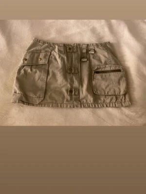 Grön cargo minikjol med fickor - Säljer en grön minikjol med flera stora fickor, dekorativa knappar och detaljer framtill och baktill. Perfekt för dig som gillar streetstyle och praktiska detaljer.