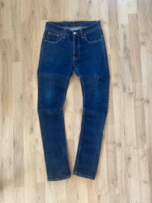 Mörkblå skinny jeans från Nudie - Snygga mörkblå skinny jeans från Nudie med klassiska orange sömmar och fem fickor. Jeansen har en smal passform och är tillverkade i stretchigt denimtyg för extra komfort. Bakfickorna har Nudies ikoniska broderi.